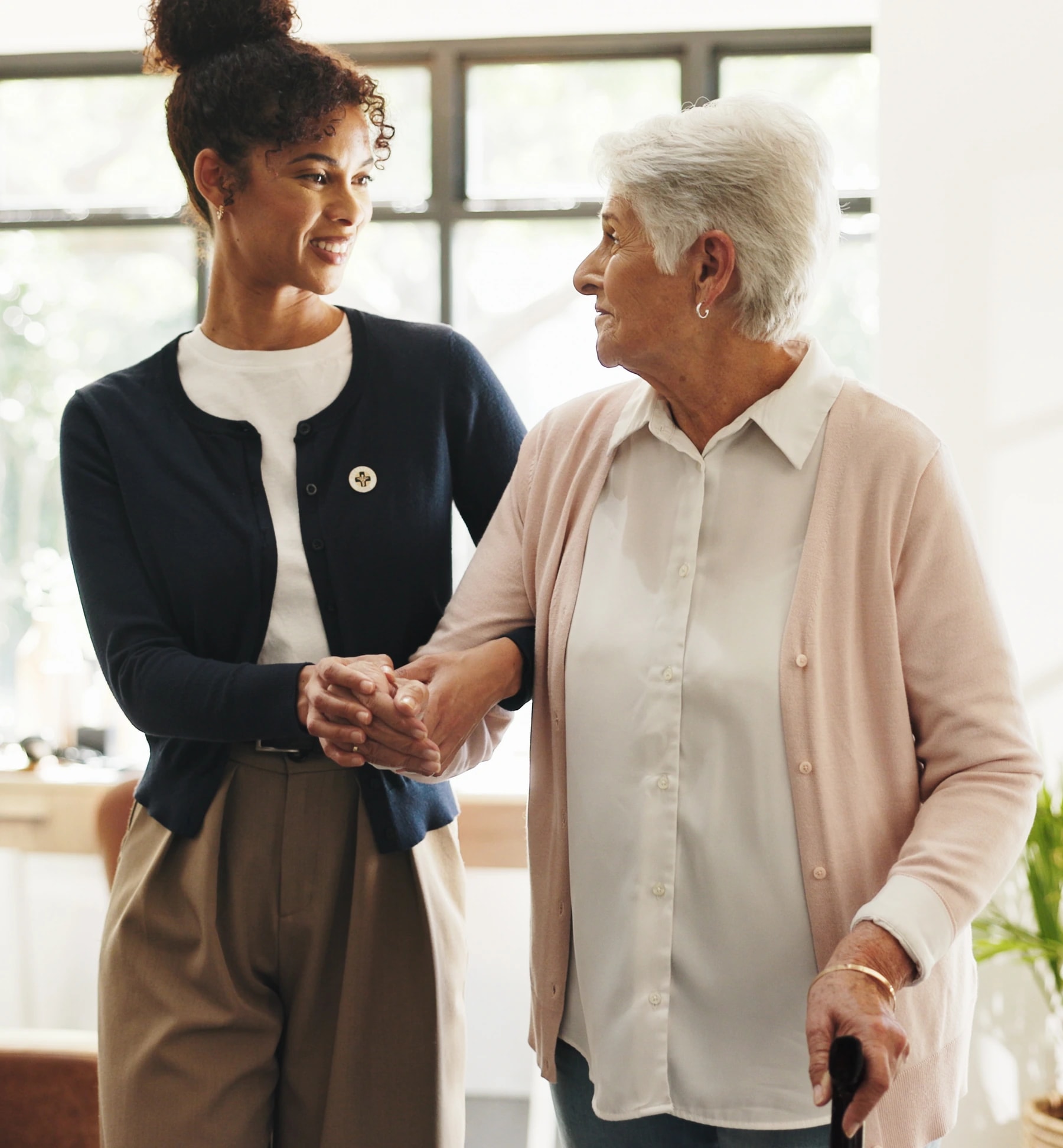 A caregiver and an older woman holding hands as they walk through a bright living room.