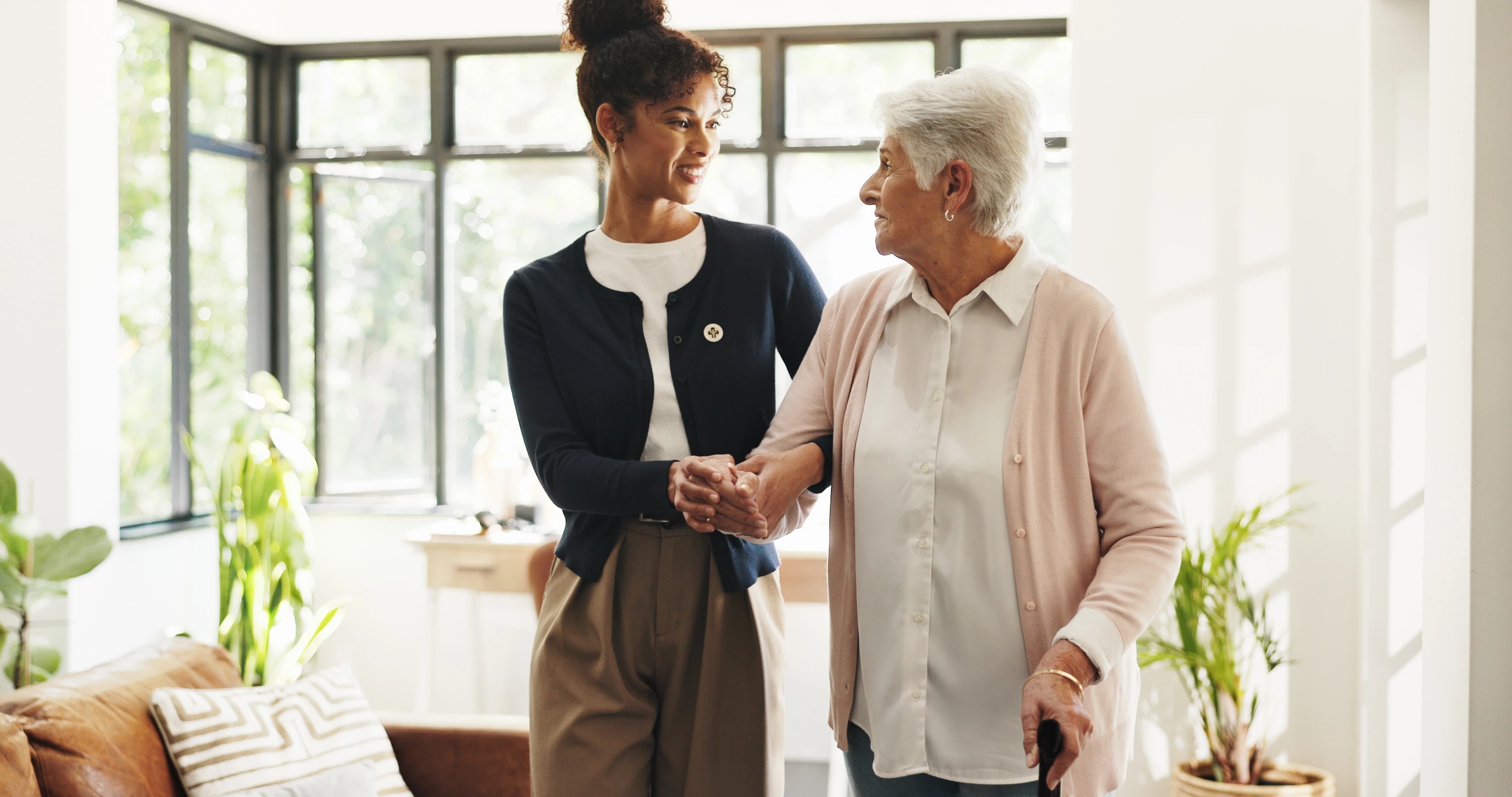 A caregiver supporting an older woman as they walk through her living room